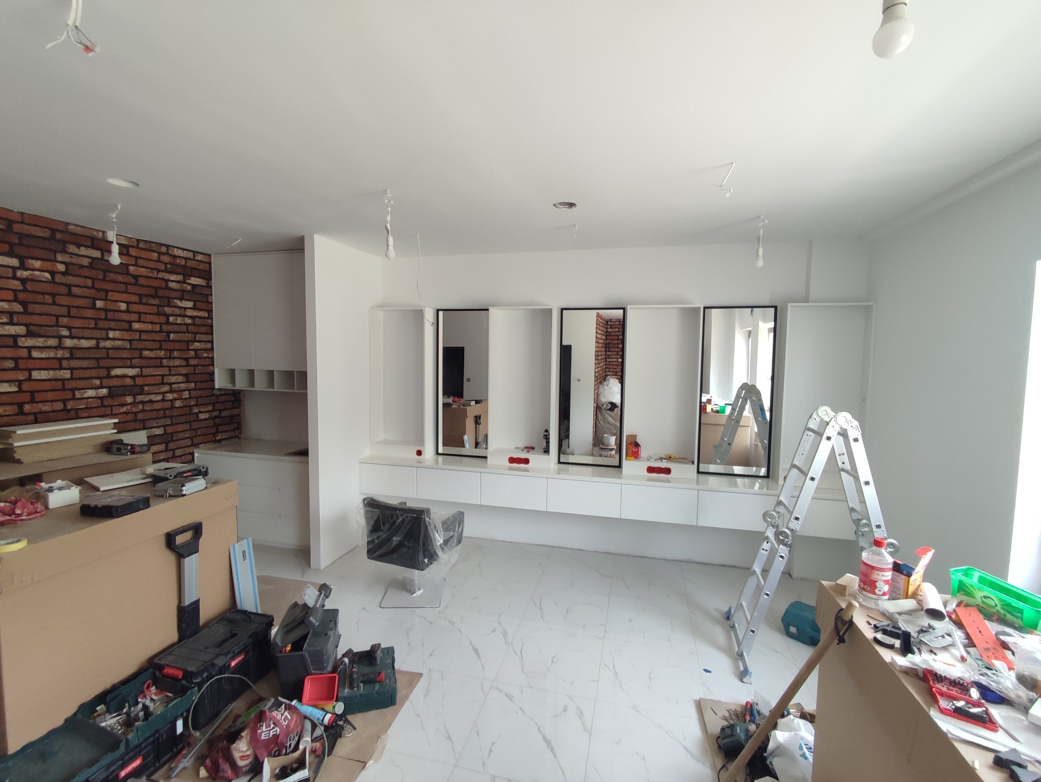 Wnętrze salonu w trakcie wykańczania z białymi szafkami pod lustrami, krzesłem fryzjerskim w folii, ceglaną ścianą, rozłożoną drabiną i narzędziami na podłodze.