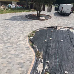 Niedokończona nawierzchnia z kostki brukowej, widoczna czarna agrowłóknina z piaskiem i przycięte pniaki, biały samochód dostawczy w tle.