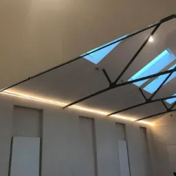 Wnętrze hali z widoczną konstrukcją stalową dachu, świetlikami i liniowym oświetleniem LED na ścianie, w tle widoczne drzwi.