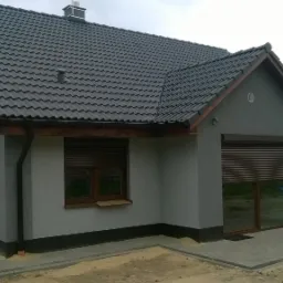 Wykończony, parterowy dom z szarą elewacją, grafitowym dachem z dachówki ceramicznej, brązowymi roletami zewnętrznymi i klimatyzacją na ścianie szczytowej.