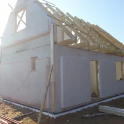 Wznoszony budynek z prefabrykatów betonowych z widoczną konstrukcją dachu z drewna, przygotowane otwory okienne i drzwiowe, na placu budowy leżą stosy drewna.