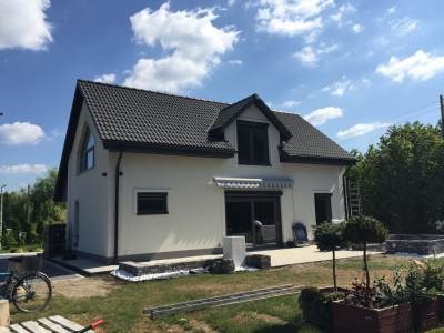 Nowoczesny, biały dom z ciemną dachówką i dużymi oknami, widok z boku w słoneczny dzień, z elementami ogrodu w trakcie aranżacji.