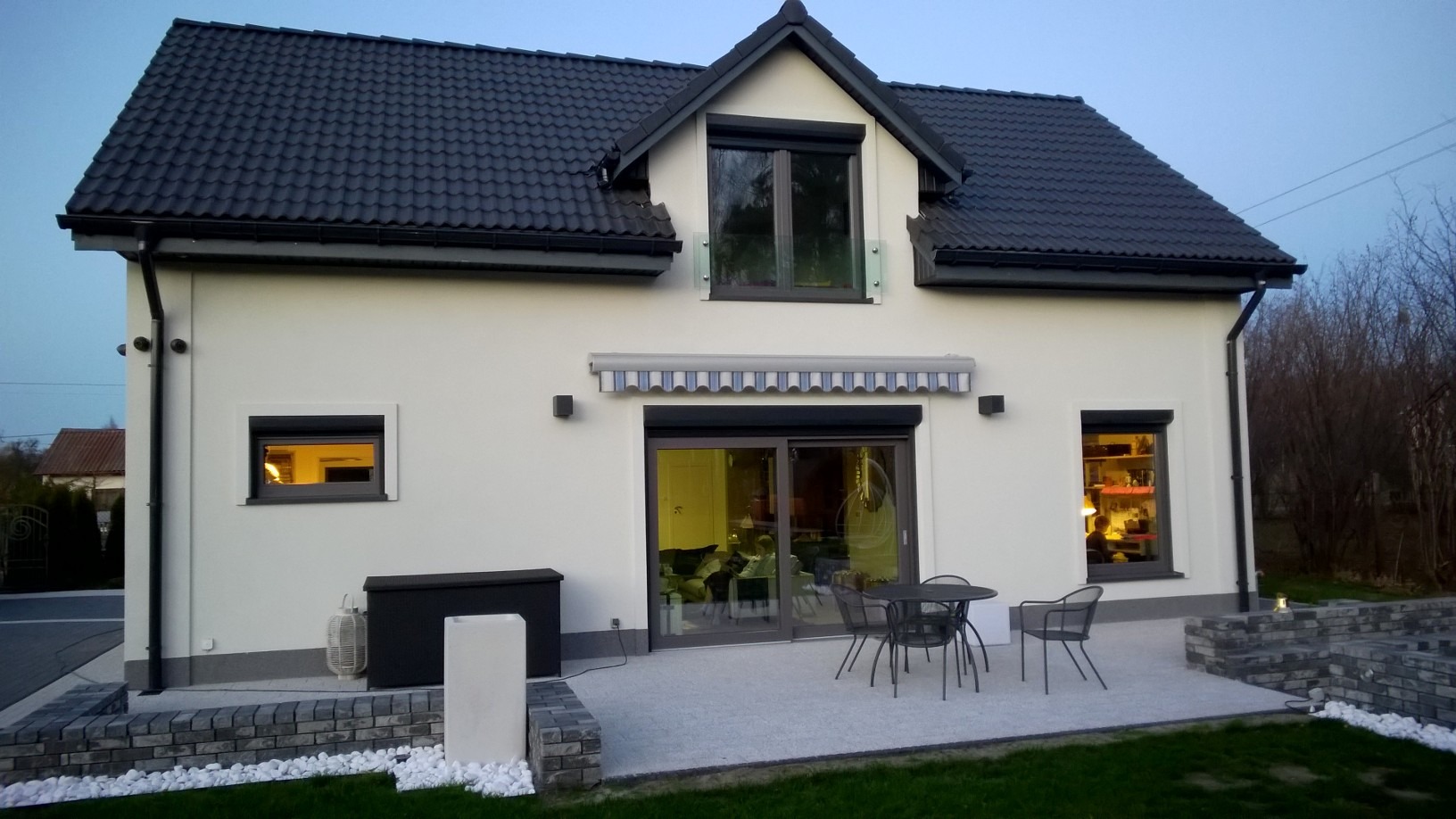 Gotowy dom szkieletowy z czarną dachówką, roletami zewnętrznymi i tarasem z szarych płyt w wieczornym oświetleniu.