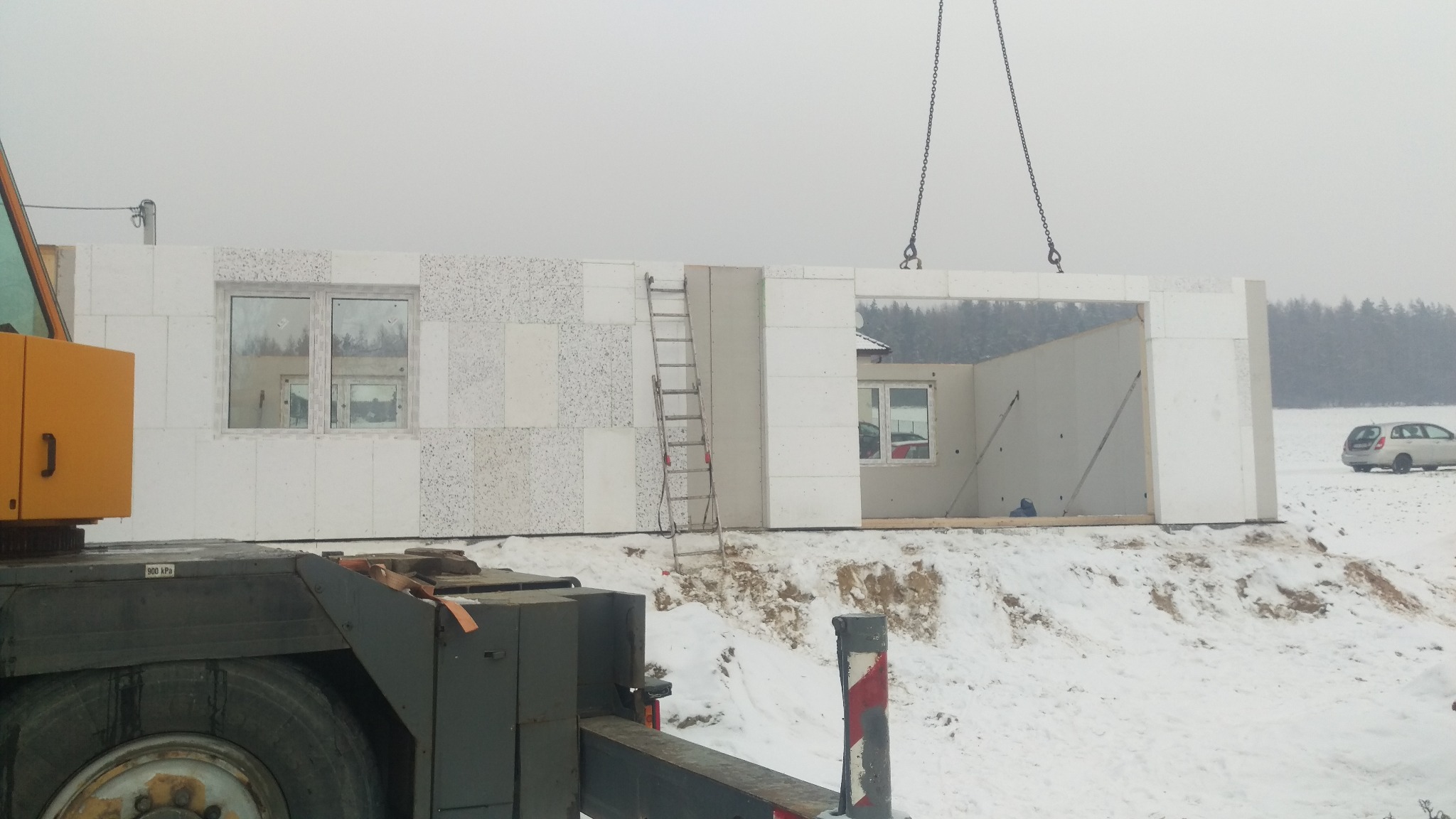 Montaż prefabrykowanej ściany z oknami na placu budowy w zimowej scenerii, widoczny dźwig i drabina, w tle zasypane śniegiem pole.