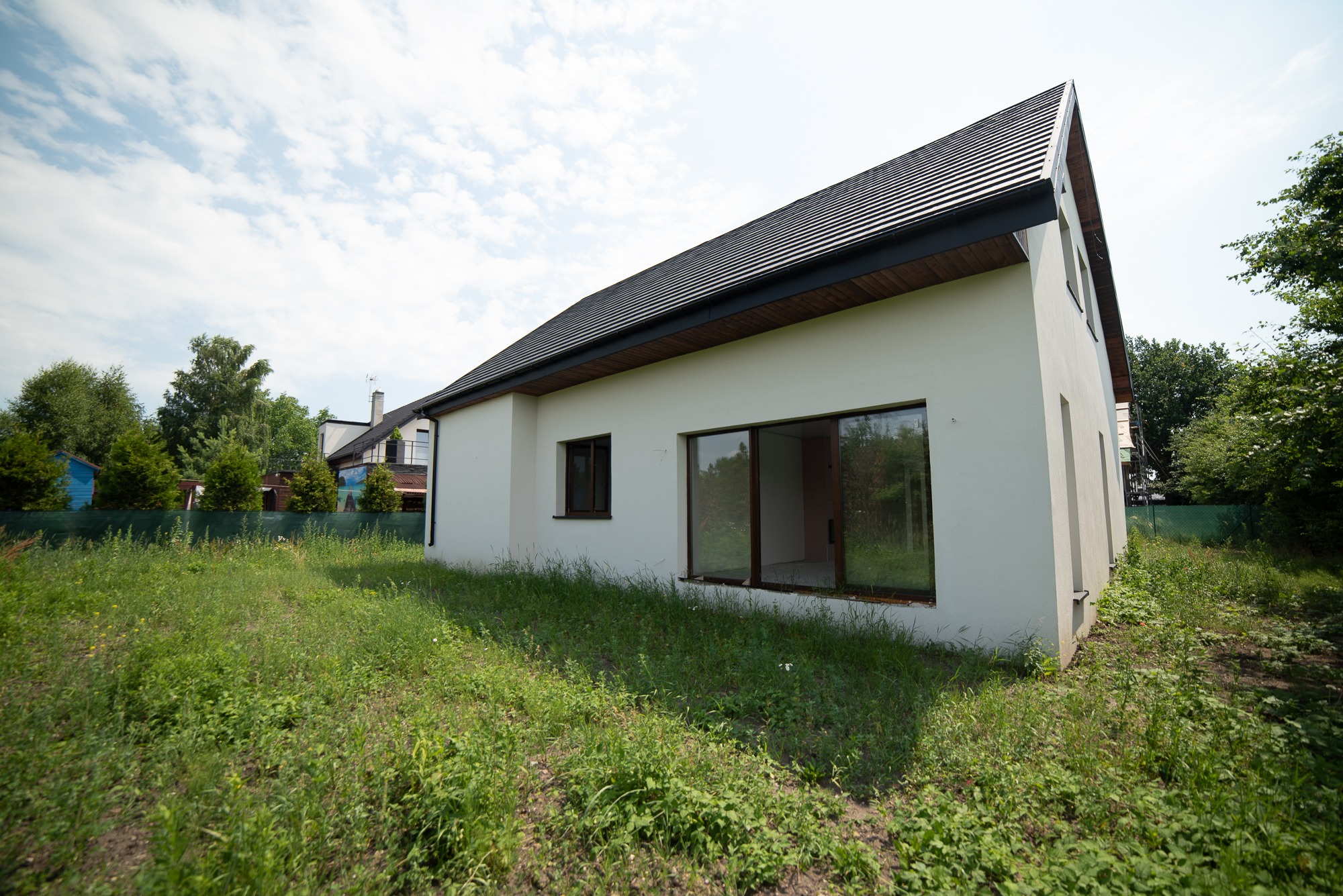 Nowoczesny, parterowy dom z białymi ścianami i ciemną dachówką, otoczony wysoką trawą i zielenią, widoczny z boku pod kątem, z oknami bez firan.