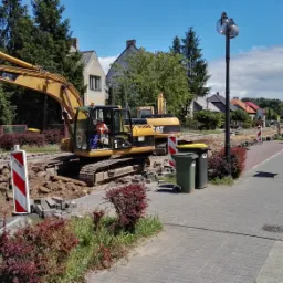 Żółta koparka CAT prowadzi prace ziemne przy chodniku z kostki brukowej, w tle zabudowa miejska i drzewa, widoczne znaki ostrzegawcze i dwa kosze na śmieci.