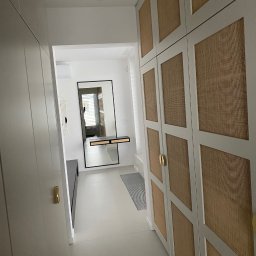 Konrad Leksinski Development - Nowoczesny przedpokój z dużym lustrem i zabudową szafy z rattanowymi frontami. Minimalistyczny design, jasne kolory, złote uchwyty. Widok z perspektywy wejścia.