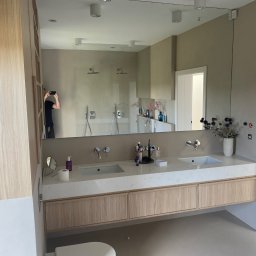 Konrad Leksinski Development - Nowoczesna łazienka z dwoma umywalkami na blacie z jasnego kamienia i drewnianymi szafkami. W lustrze odbija się kabina prysznicowa i osoba robiąca zdjęcie.