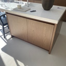 Konrad Leksinski Development - Nowoczesna wyspa kuchenna z drewnianymi frontami i białym blatem. Na blacie wazon, szklanki i dekoracje. Jasne, minimalistyczne wnętrze.