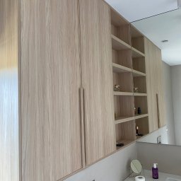 Konrad Leksinski Development - Jasne, drewniane szafki łazienkowe z półkami, widok z dołu, minimalistyczny design, lustro odbijające przestrzeń. Nowoczesne wykończenie.