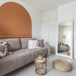 Konrad Leksinski Development - Jasny salon z beżową kanapą, okrągłym stolikiem i lustrem. Na ścianie pomalowany łuk w kolorze terakoty. Widoczne białe listwy dekoracyjne i białe meble.