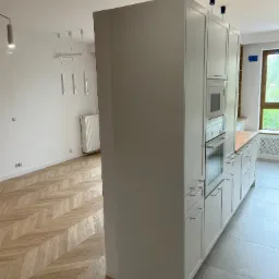 Nowoczesna kuchnia z białą zabudową, drewnianym blatem i parkietem w jodełkę. Widoczny sprzęt AGD w zabudowie i szara podłoga. Minimalistyczny design.