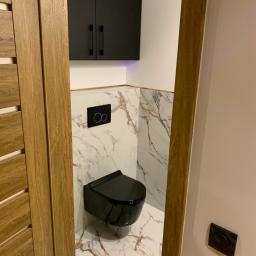 Konrad Leksinski Development - Wykończona łazienka z wiszącą, czarną miską WC, marmurowymi płytkami z żyłkowaniem w kolorze złota, czarną szafką nad WC i drewnianymi drzwiami.