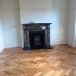 Konrad Leksinski Development - Pokój po remoncie z parkietem w jodełkę i ozdobnym kominkiem z czarnym wkładem.