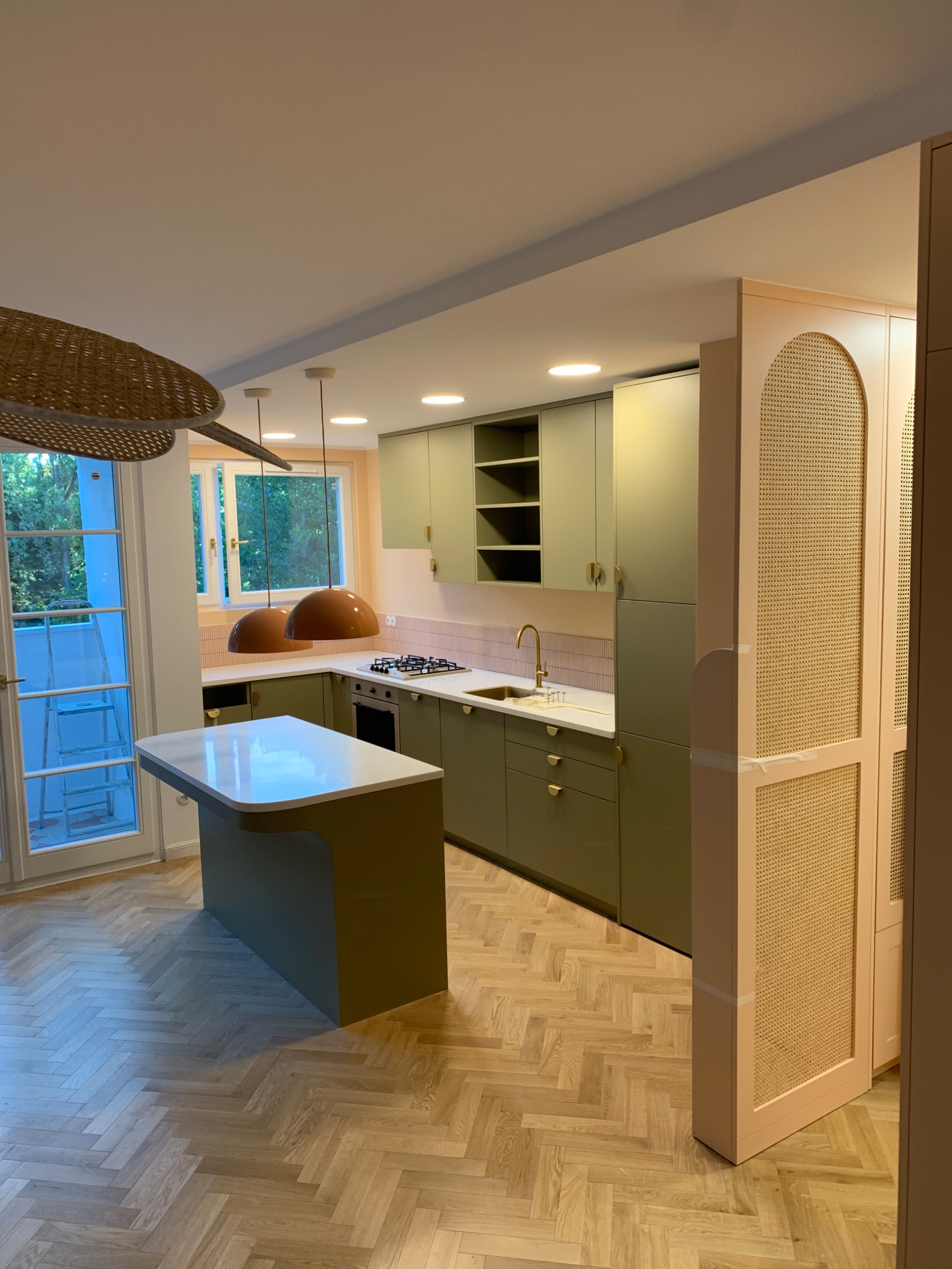 Nowoczesna kuchnia w odcieniach zieleni i beżu, z wyspą i parkietem w jodełkę. Widoczne lampy wiszące i drzwi z rattanowym wypełnieniem.