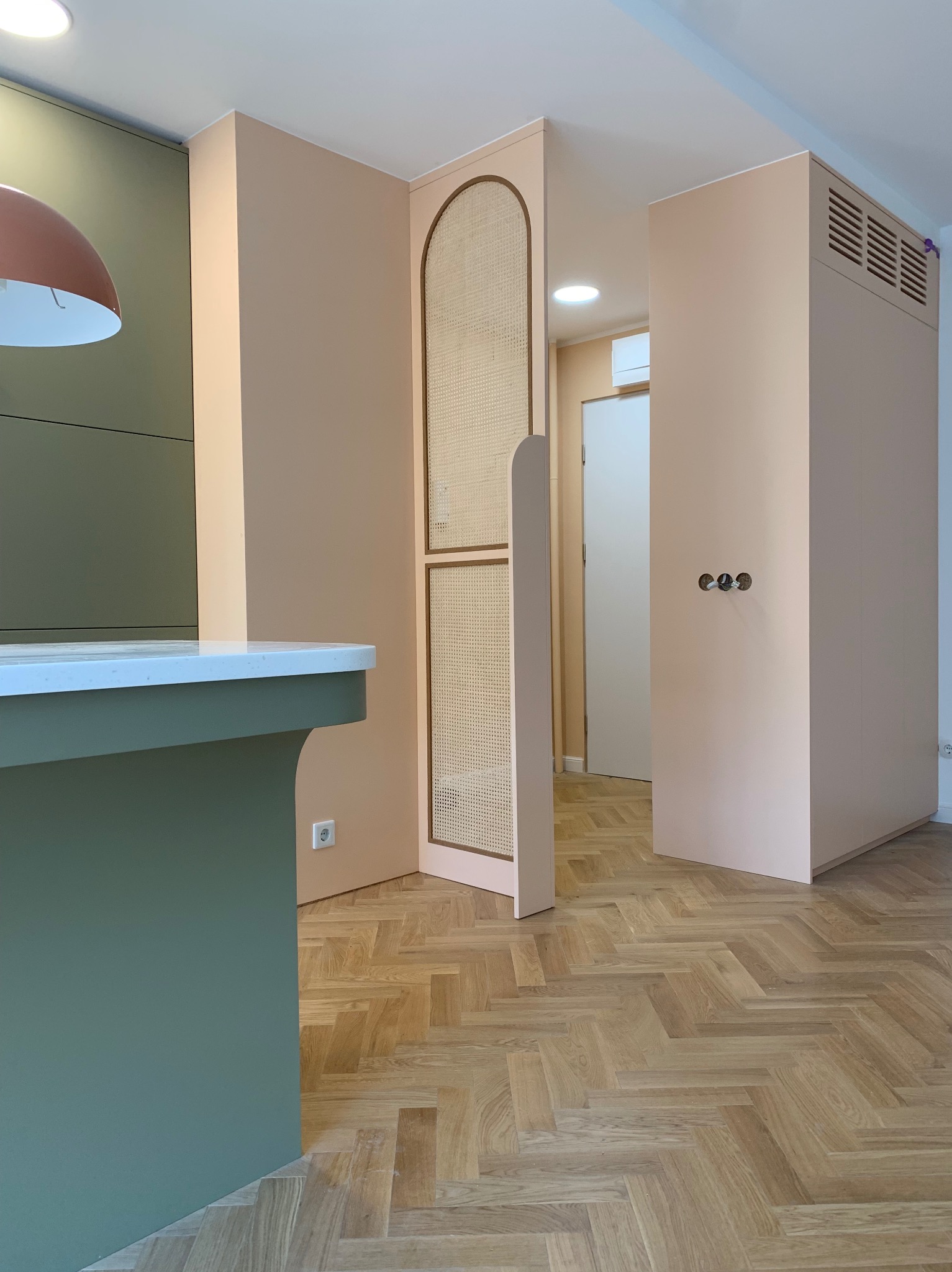 Nowoczesne wykończenie wnętrza: parkiet w jodełkę, zabudowa w odcieniach beżu i zieleni, dekoracyjna ścianka z rattanem, minimalistyczne oświetlenie.