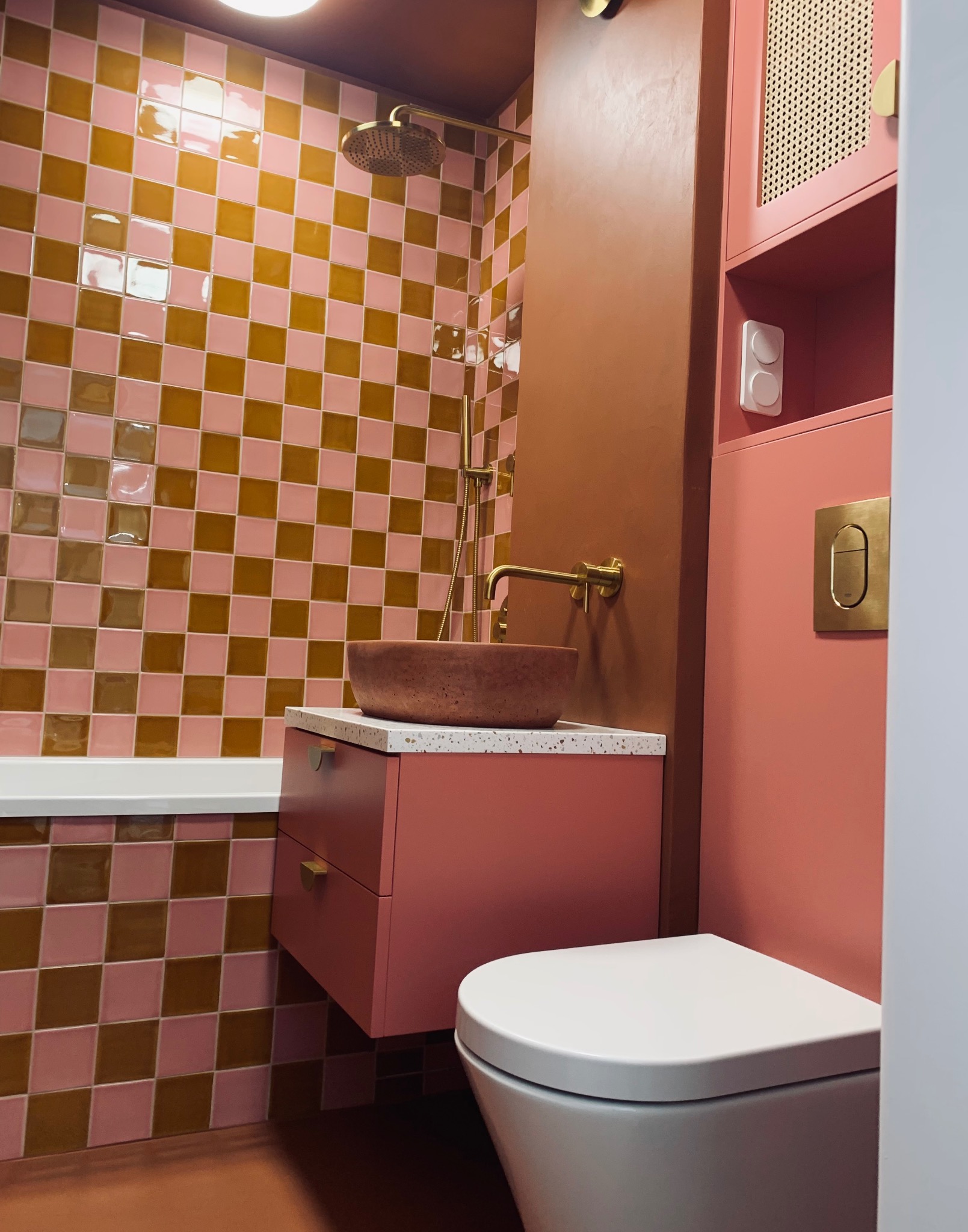 Nowoczesna łazienka w odcieniach różu i brązu. Uwagę zwracają złote detale armatury i nietypowa umywalka z kamienia. Na ścianach wzór szachownicy z płytek.