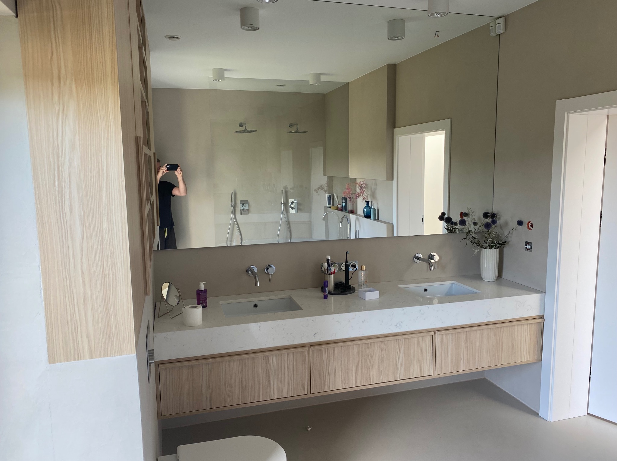 Nowoczesna łazienka z dwoma umywalkami na blacie z jasnego kamienia i drewnianymi szafkami. W lustrze odbija się kabina prysznicowa i osoba robiąca zdjęcie.