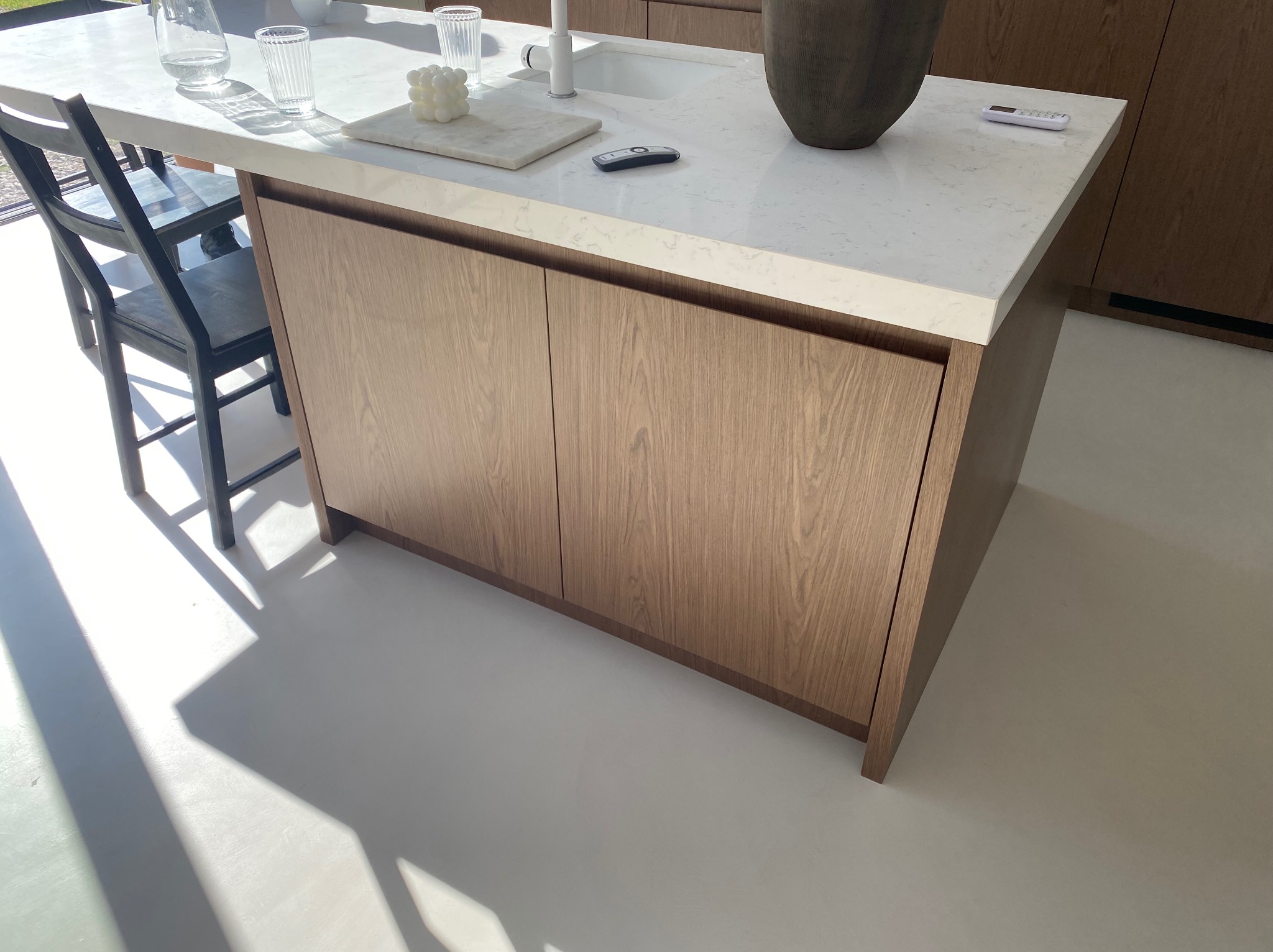 Nowoczesna wyspa kuchenna z drewnianymi frontami i białym blatem. Na blacie wazon, szklanki i dekoracje. Jasne, minimalistyczne wnętrze.