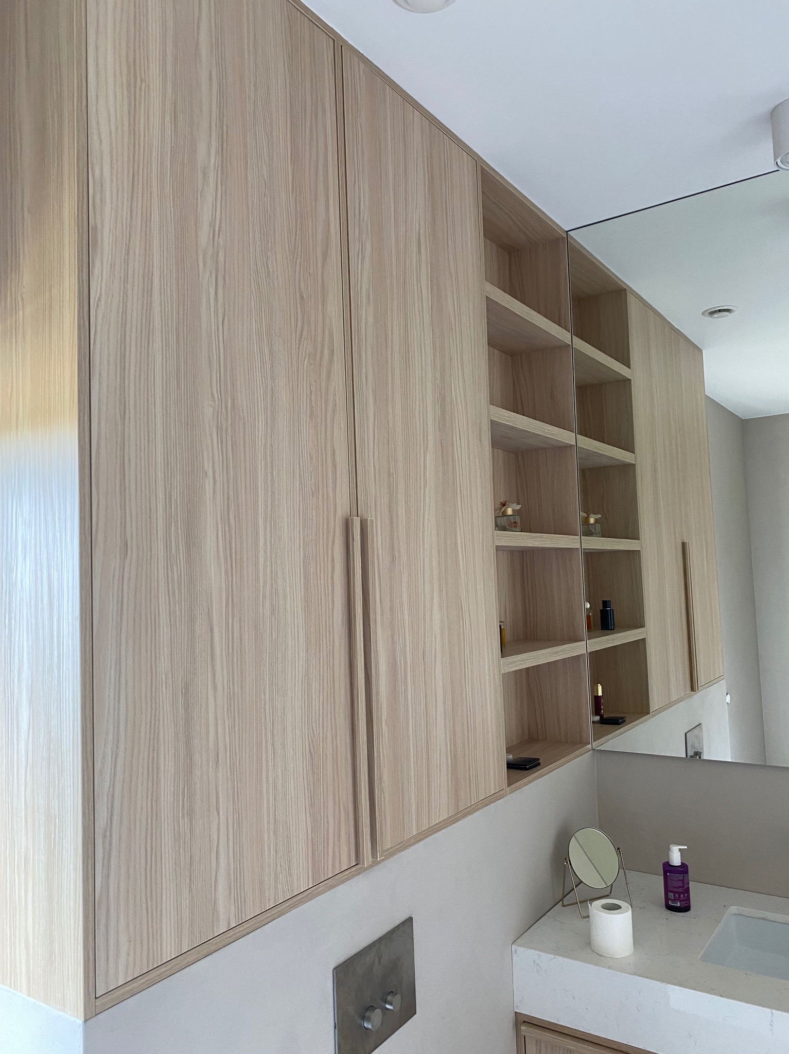 Jasne, drewniane szafki łazienkowe z półkami, widok z dołu, minimalistyczny design, lustro odbijające przestrzeń. Nowoczesne wykończenie.