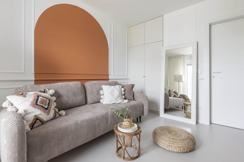 Jasny salon z beżową kanapą, okrągłym stolikiem i lustrem. Na ścianie pomalowany łuk w kolorze terakoty. Widoczne białe listwy dekoracyjne i białe meble.