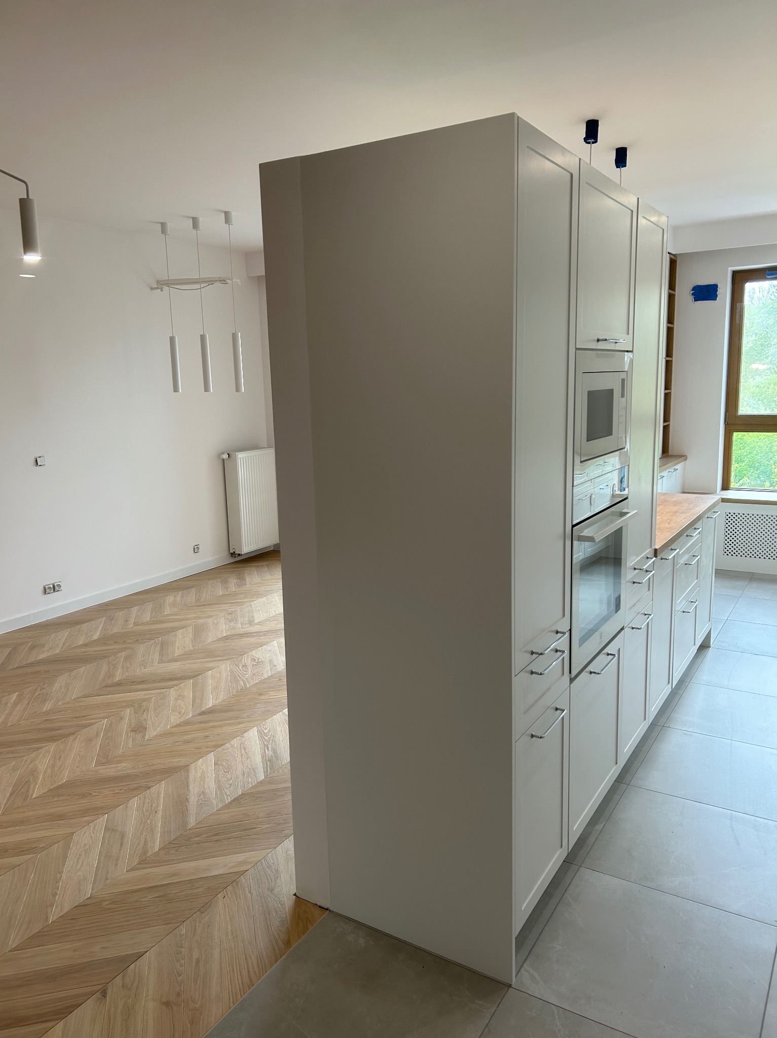 Nowoczesna kuchnia z białą zabudową, drewnianym blatem i parkietem w jodełkę. Widoczny sprzęt AGD w zabudowie i szara podłoga. Minimalistyczny design.