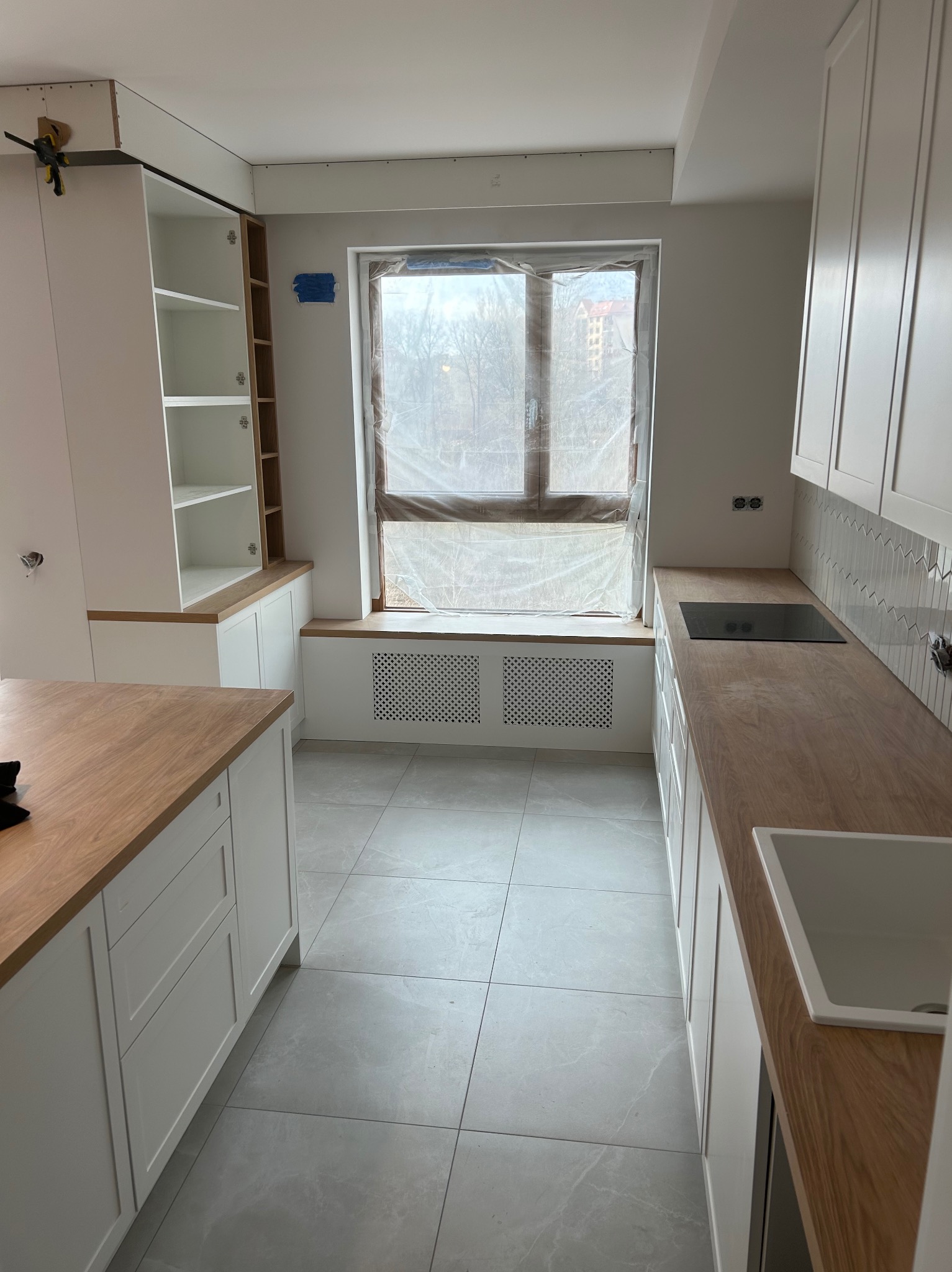 Jasna kuchnia w trakcie montażu: białe szafki z drewnianym blatem, okno zabezpieczone folią. Widoczne niedokończone elementy i szare płytki podłogowe.
