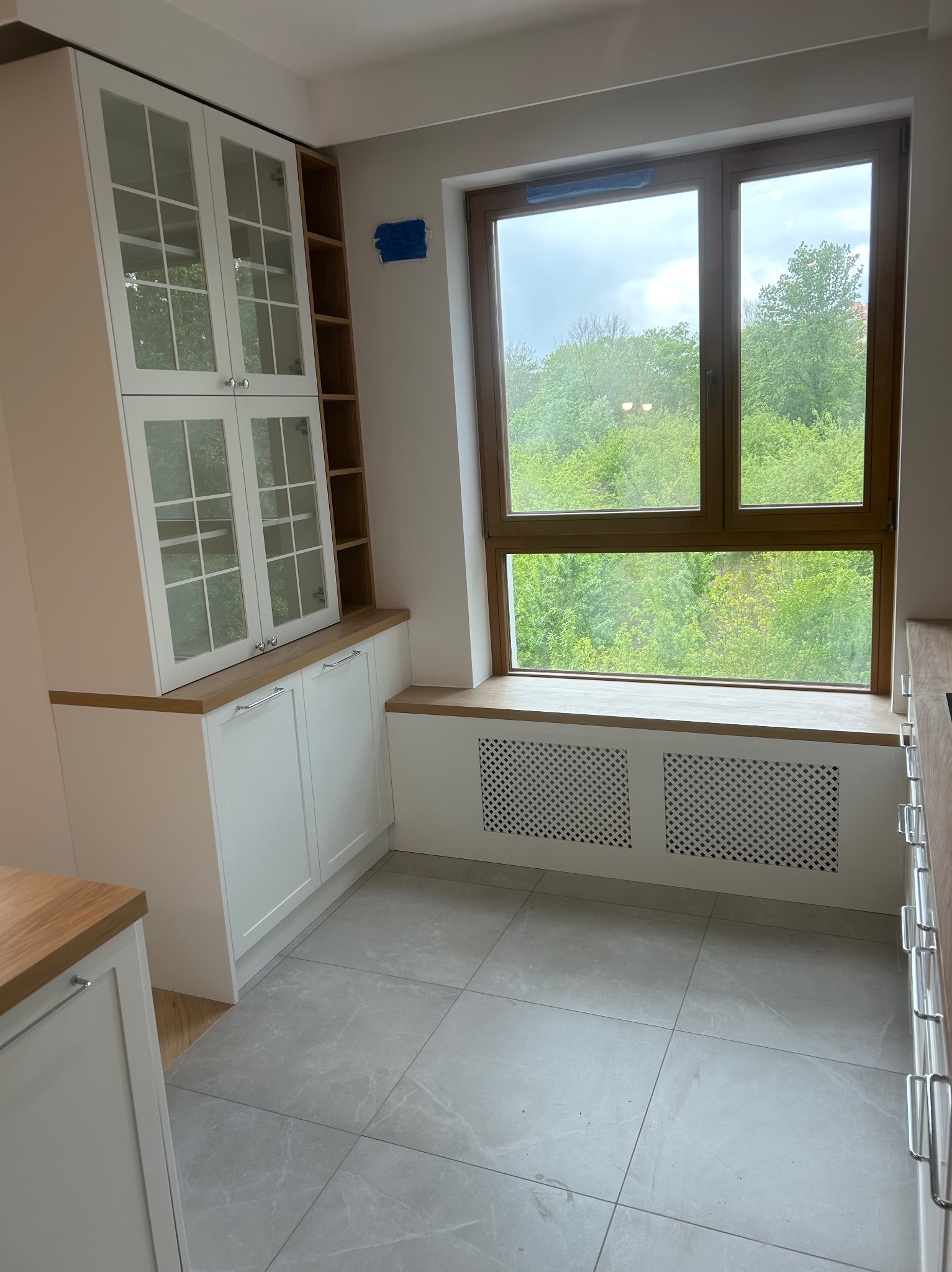Jasna kuchnia z zabudową okienną i szafkami z przeszkleniami. Widok na zieleń za oknem. Szare płytki podłogowe.