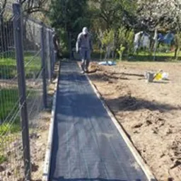 Przygotowanie podłoża pod trawnik z rolki: dwóch mężczyzn w roboczych ubraniach rozkłada czarną agrowłókninę na wytyczonym pasie ziemi, obramowanym drewnianymi deskami. W tle widoczne drzewa...