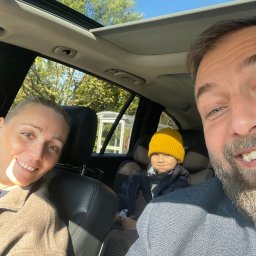 PGH - Rodzinne selfie w samochodzie z otwartym szyberdachem, widoczne drzewa i błękitne niebo, dziecko w żółtej czapce siedzące w foteliku samochodowym.