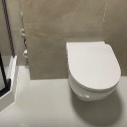 PGH - Nowoczesna, podwieszana toaleta w wykończonej łazience z beżowymi płytkami i czarnym panelem sterującym spłuczką.