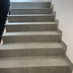 PGH - Schody wyłożone szarymi płytkami ceramicznymi, z widocznymi fugami, czarna metalowa barierka po lewej stronie, ściana pomalowana na biało, widoczne drobne zabrudzenia po pracach wykończeniowych.