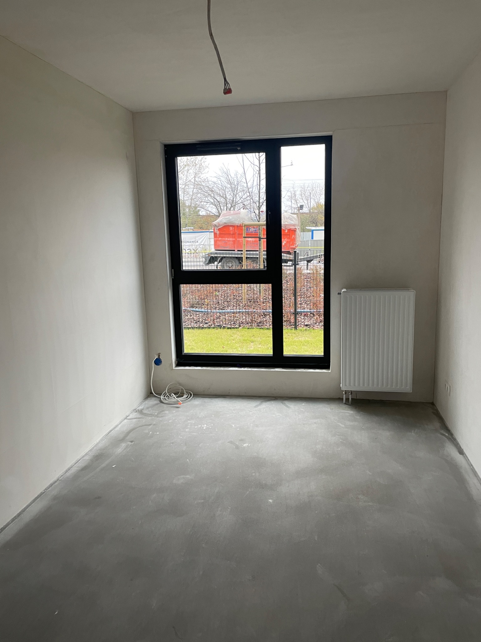 Pokój w stanie deweloperskim: surowe ściany, wylewka betonowa na podłodze, grzejnik panelowy pod oknem z widokiem na zewnątrz; przewód elektryczny zwisający z sufitu.