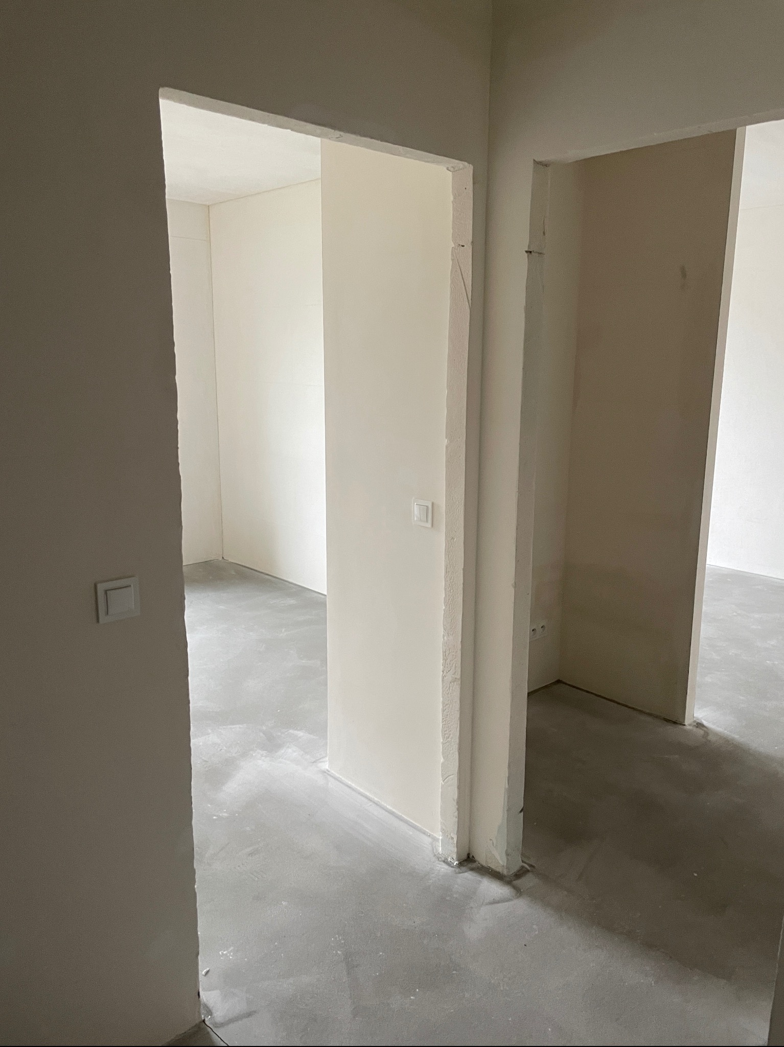 Surowe, jasne wnętrze w trakcie prac wykończeniowych; widoczne surowe ściany, wylewka podłogowa i ościeżnice z widocznymi przewodami elektrycznymi i puszkami w ścianach.