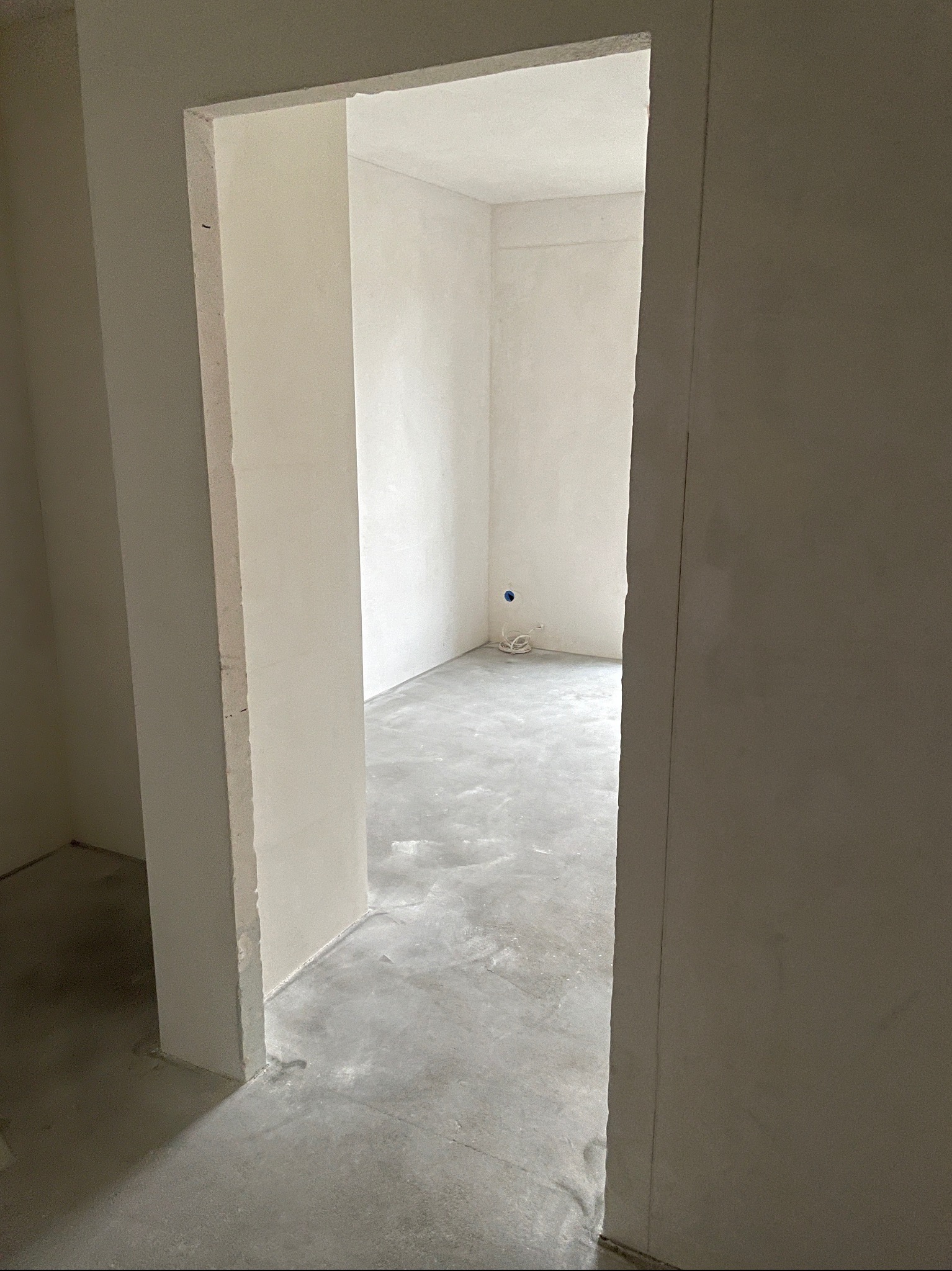 Surowy stan wnętrza z widoczną wnęką drzwiową, szpachlowane ściany, wylewka betonowa na podłodze, przewody elektryczne wystające ze ściany w głębi pomieszczenia.