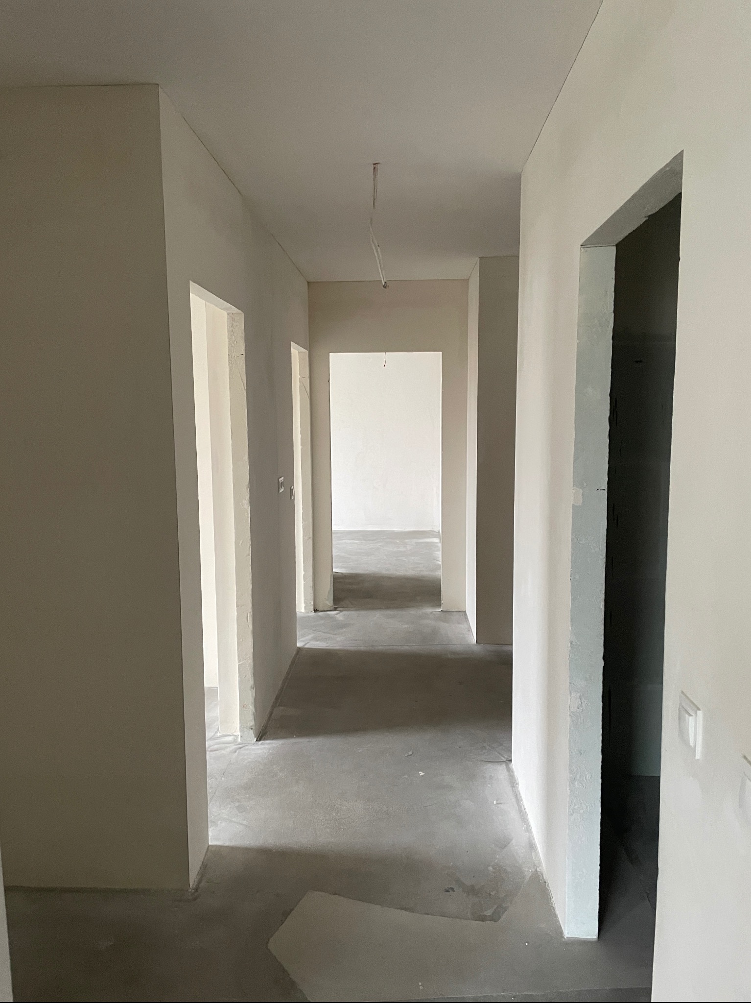 Korytarz w stanie deweloperskim, ściany pokryte gładzią, surowa wylewka na podłodze, widoczne otwory drzwiowe bez ościeżnic, przewody elektryczne wystające z sufitu.