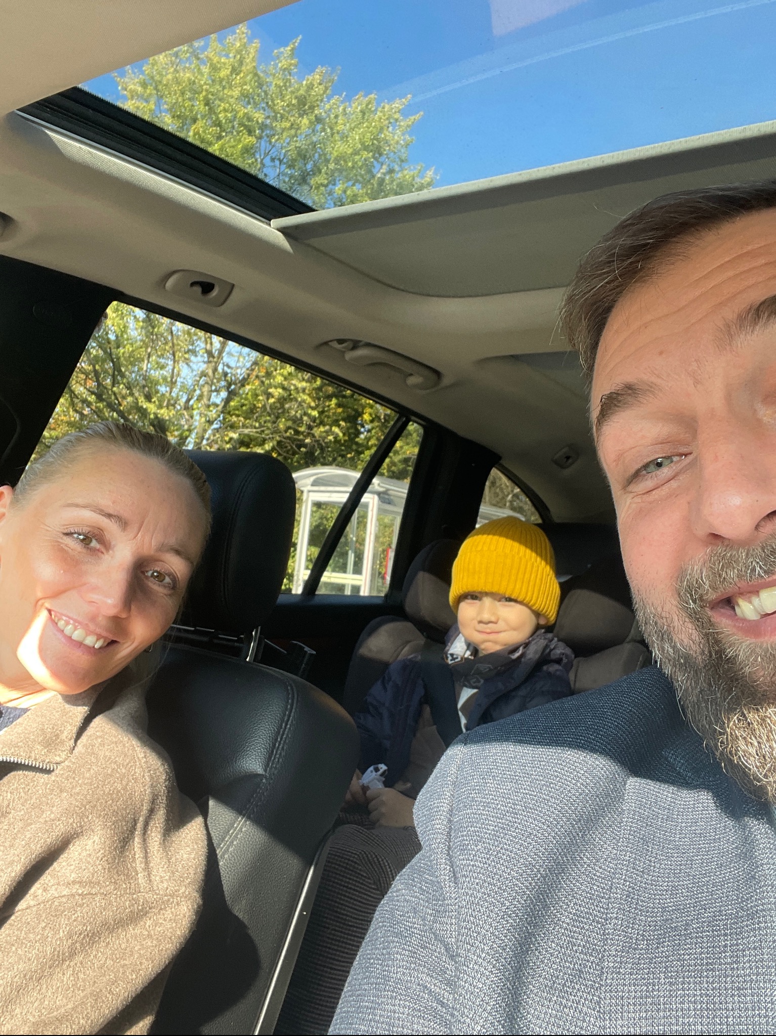 Rodzinne selfie w samochodzie z otwartym szyberdachem, widoczne drzewa i błękitne niebo, dziecko w żółtej czapce siedzące w foteliku samochodowym.