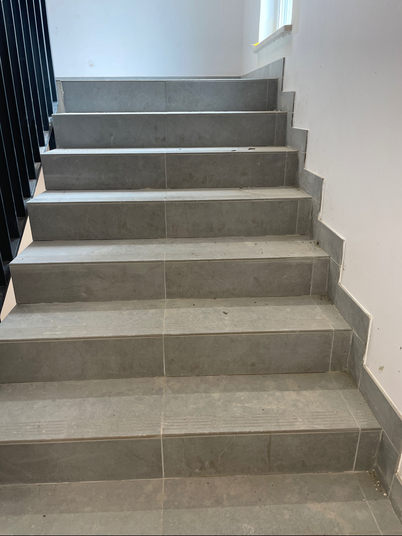 Schody wyłożone szarymi płytkami ceramicznymi, z widocznymi fugami, czarna metalowa barierka po lewej stronie, ściana pomalowana na biało, widoczne drobne zabrudzenia po pracach wykończeniowych.