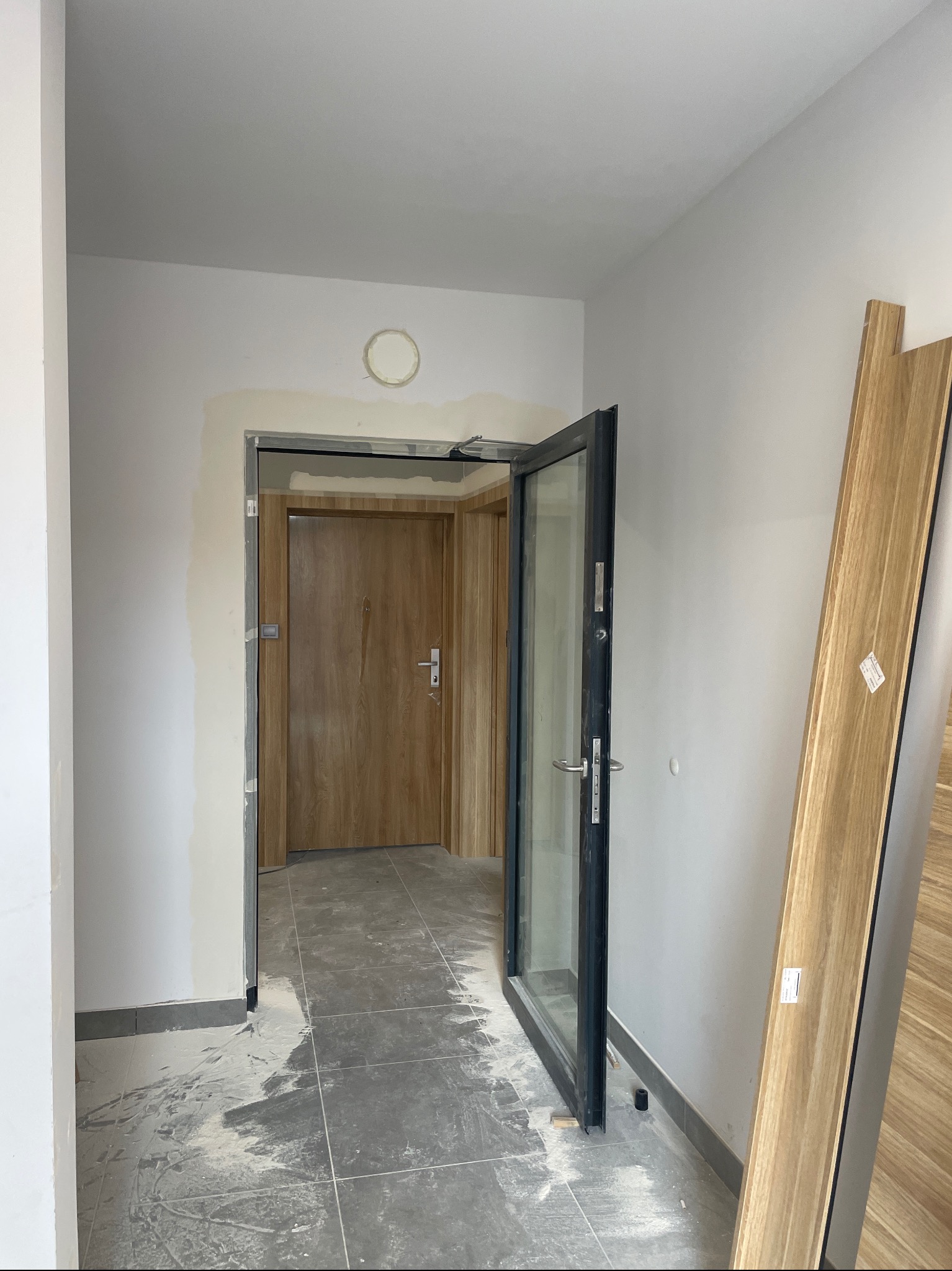 Korytarz w trakcie prac wykończeniowych z otwartymi drzwiami wejściowymi z czarną ramą i szklanym panelem oraz drewnianymi panelami drzwiowymi opartymi o ścianę, widoczne ślady po pracach na podłodze.