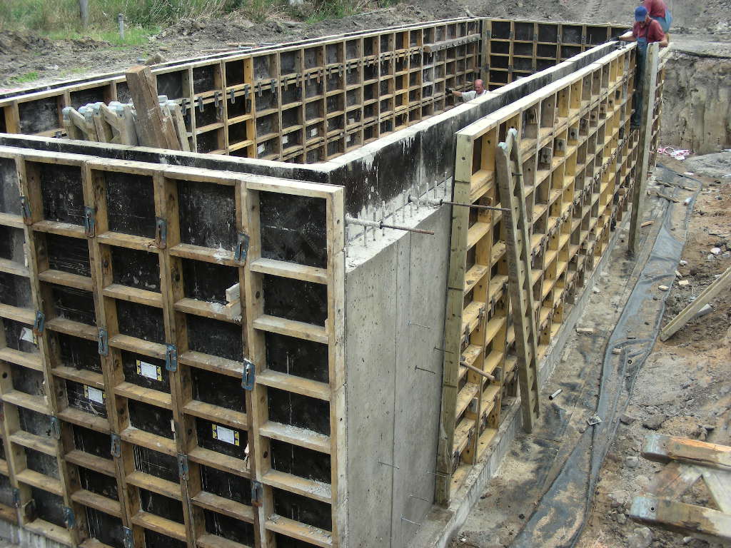 Rozbudowane szalunki drewniane tworzące formę dla wylewania fundamentów, z widocznymi elementami zbrojenia i pracownikami budowlanymi w tle.