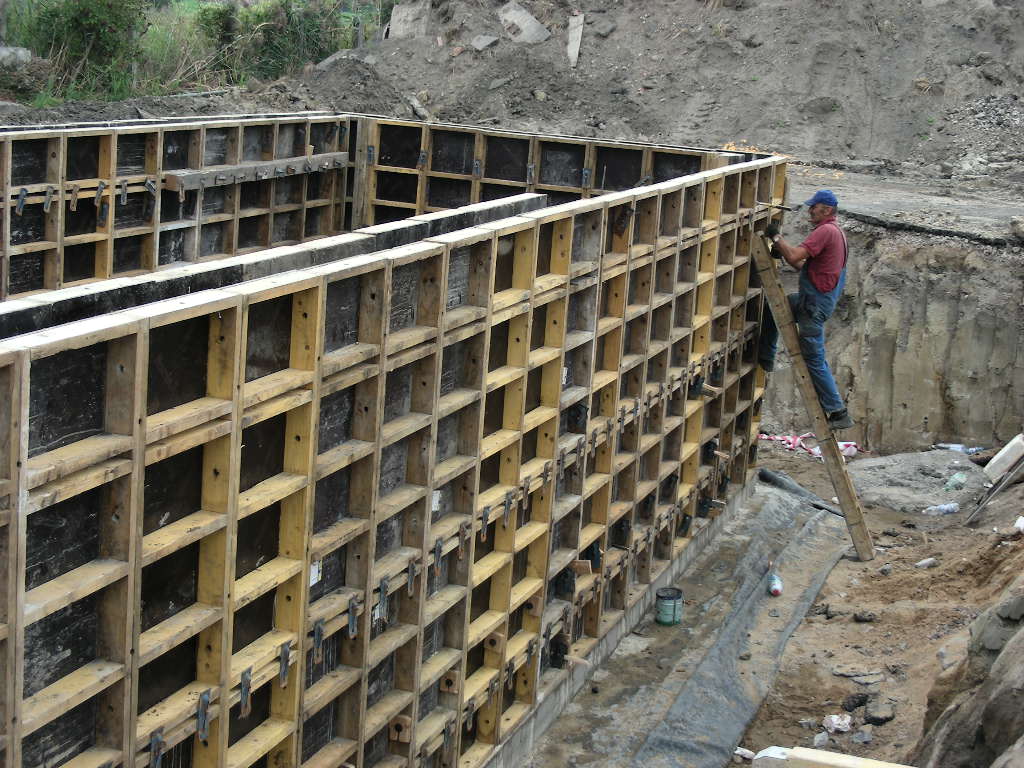 Montaż szalunków z drewnianych belek na budowie fundamentów, pracownik na drabinie przytwierdza elementy.