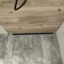 Rafal - Szare płytki podłogowe w kabinie prysznicowej z odpływem liniowym i płytki imitujące drewno na ścianie, widoczny fragment rury odpływowej.