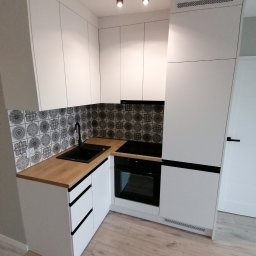 Rafal - Nowoczesna kuchnia w kształcie litery L z białymi szafkami, drewnianym blatem i czarną armaturą, wykończona płytkami z geometrycznym wzorem na ścianie nad blatem.