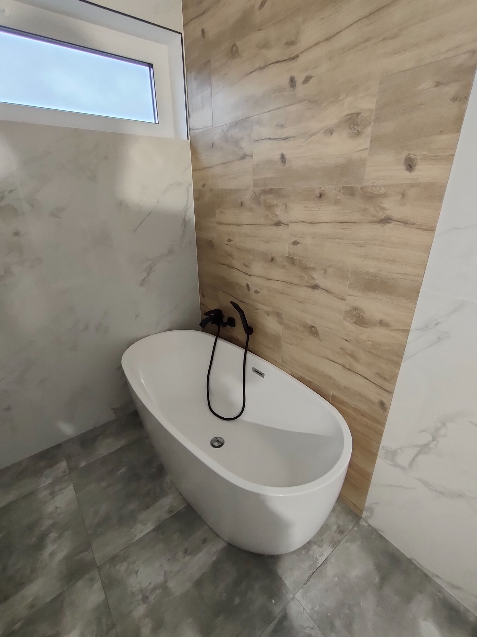 Nowoczesna łazienka z wolnostojącą białą wanną, czarną armaturą, płytkami imitującymi drewno na ścianie i marmurowymi płytkami na obudowie wanny oraz szarą podłogą.