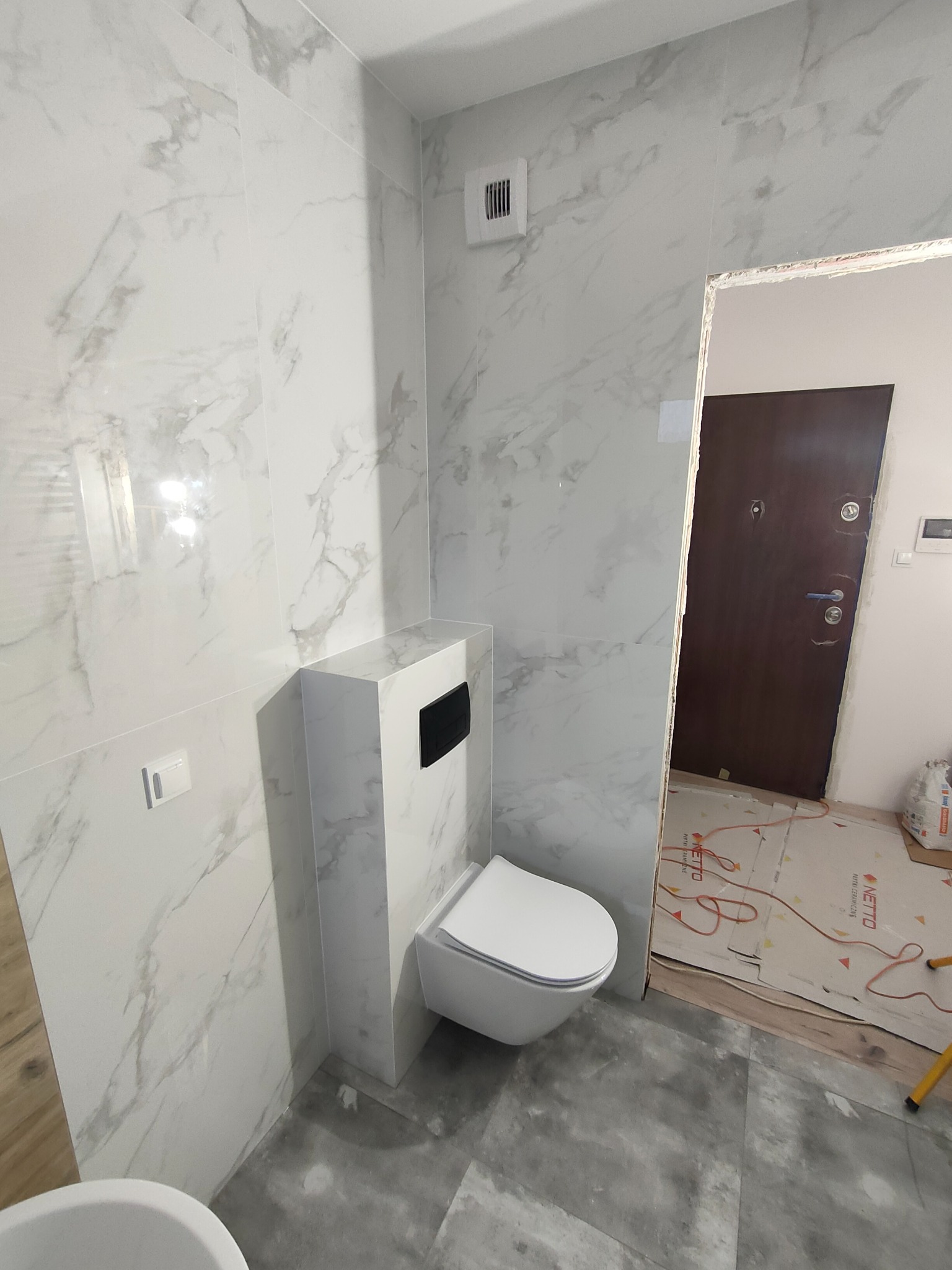 Wnętrze łazienki z toaletą wiszącą na tle marmurowych płytek ściennych i szarych płytek podłogowych, widoczny fragment kartonów ochronnych przy wejściu.