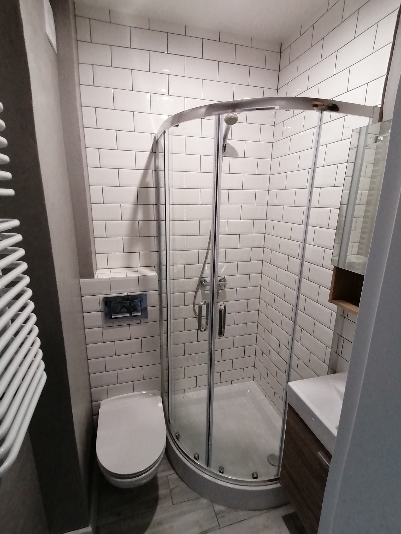 Nowoczesna łazienka z kabiną prysznicową narożną, białą armaturą i płytkami typu metro na ścianach, uzupełniona grzejnikiem drabinkowym i szafką z umywalką w odcieniach brązu.