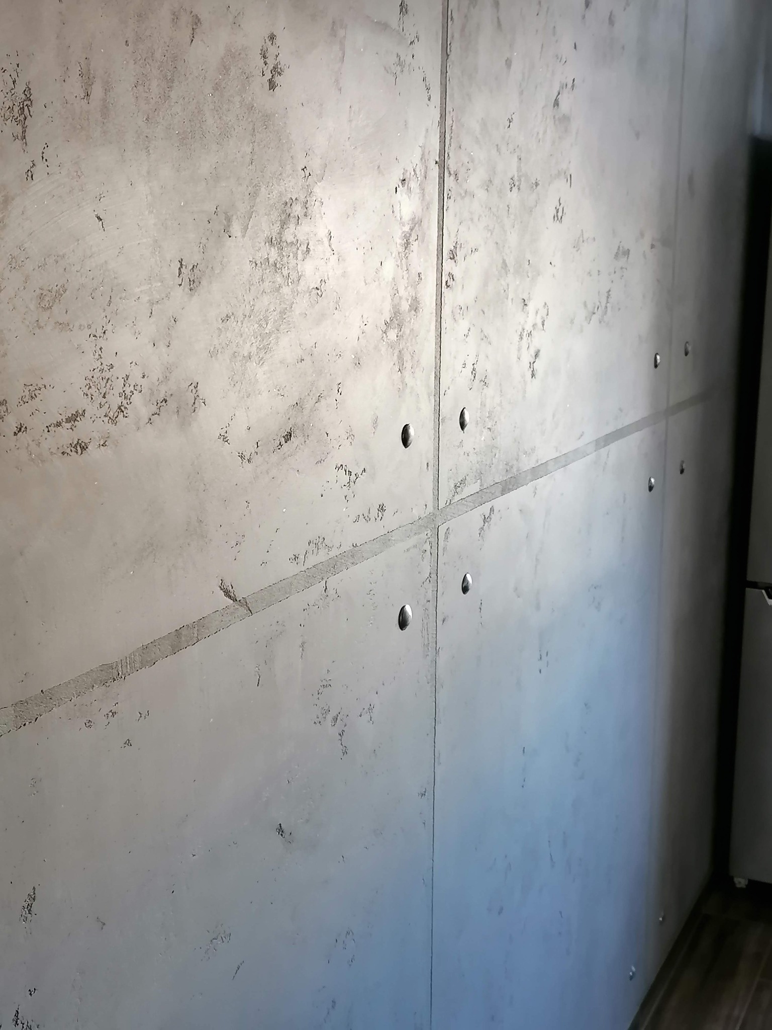 Szara ściana wykończona panelami imitującymi beton, połączonymi srebrnymi nitami, z widoczną fakturą i nierównomiernym odcieniem szarości.