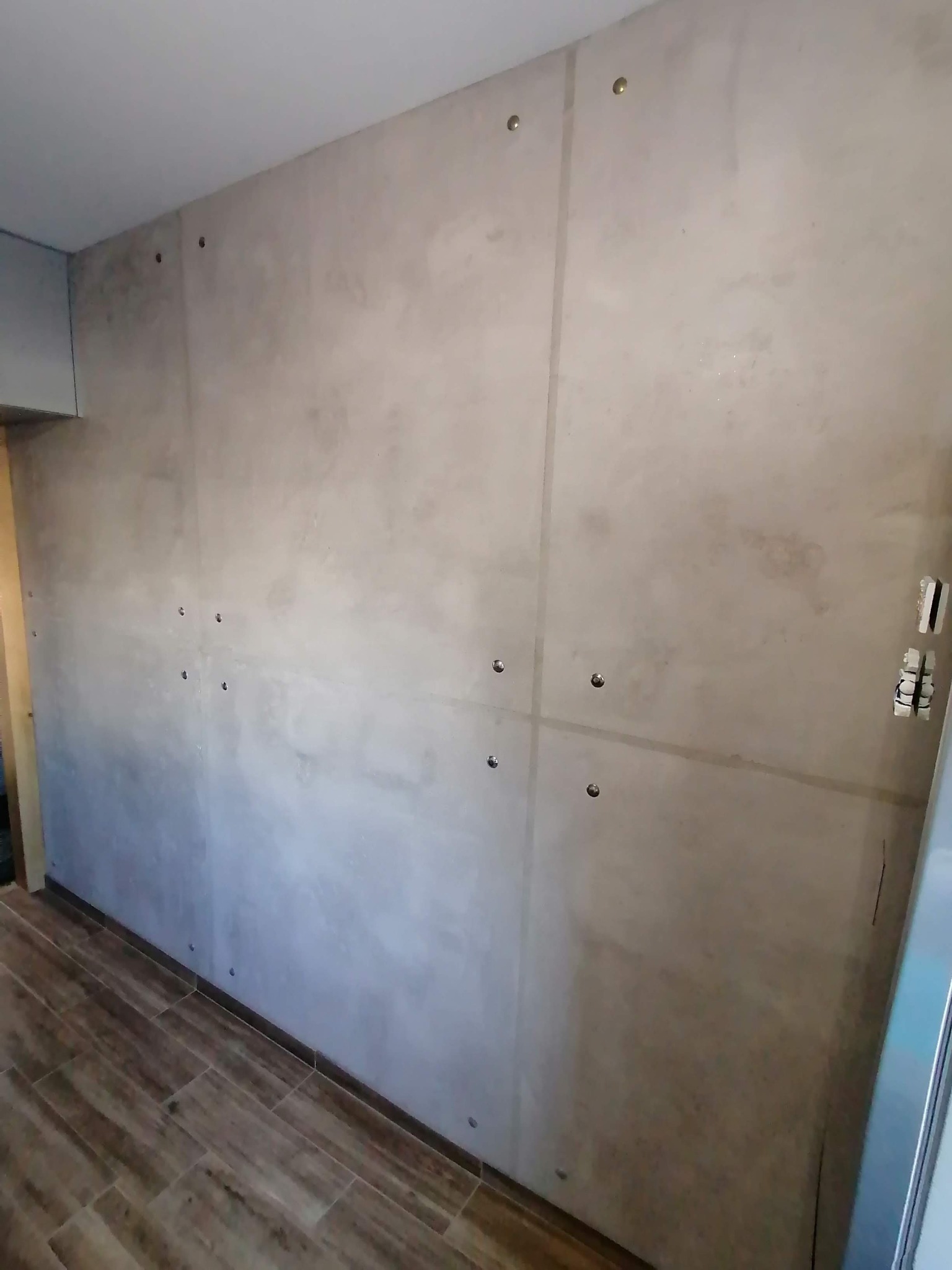 Ściana wykończona szarym betonem architektonicznym z widocznymi otworami montażowymi i delikatnymi pionowymi podziałami, brązowa podłoga z płytek imitujących drewno.