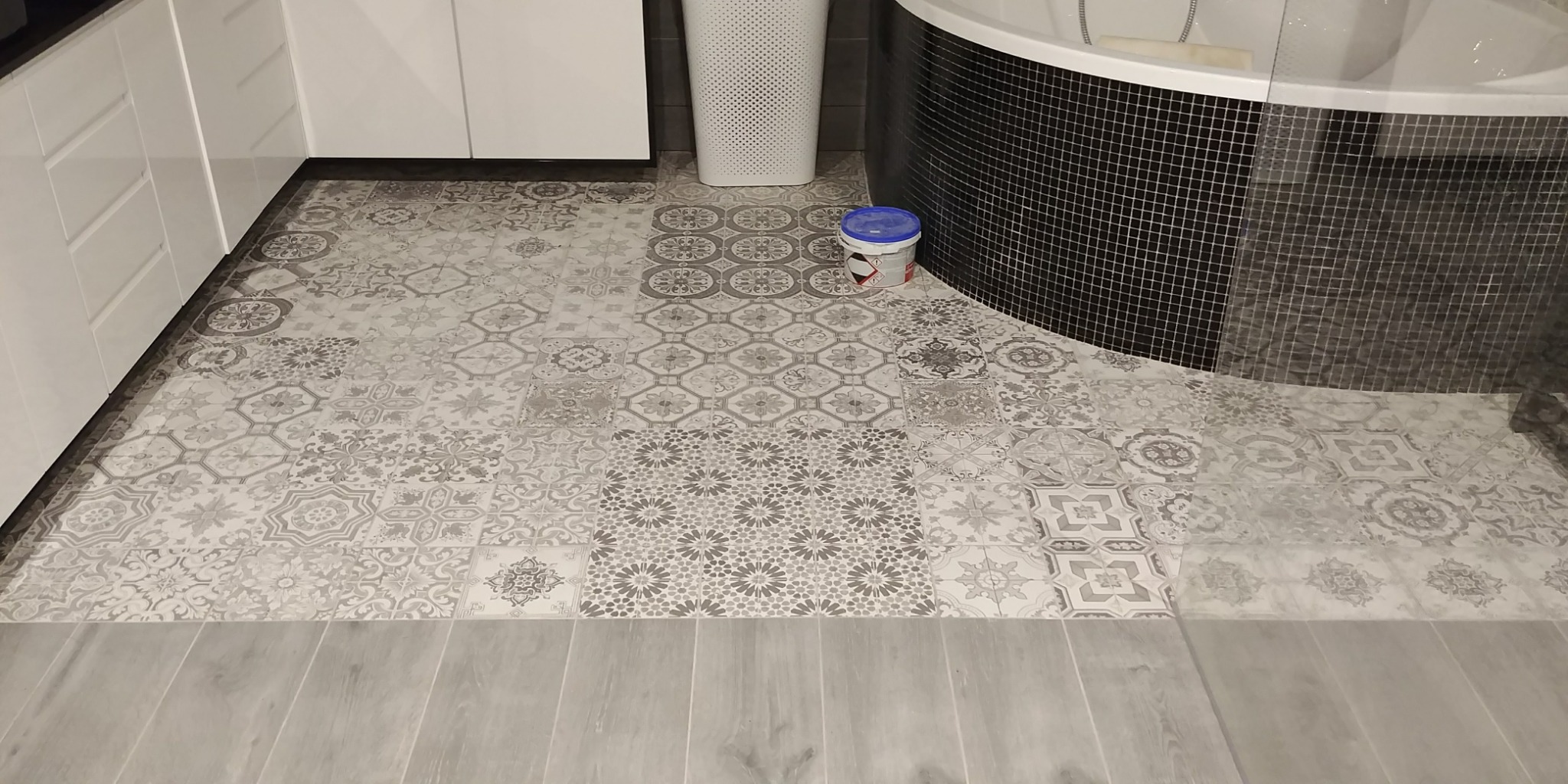 Ukończona łazienka z mozaikową ścianą wanny i patchworkową podłogą z płytek, widoczny pojemnik z niebieskim wieczkiem po prawej stronie.