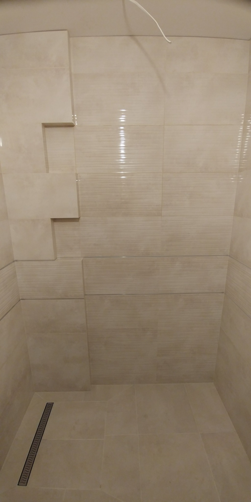 Wykończona kabina prysznicowa z beżowymi płytkami o zróżnicowanej fakturze, z odpływem liniowym w podłodze i półkami wbudowanymi w ścianę.